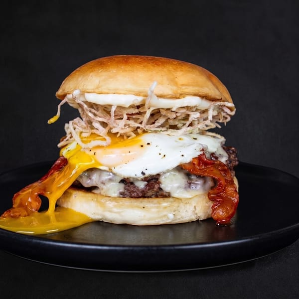 Gourmet burger with a runny fried egg, crispy bacon, melted cheese, and shredded toppings on a black plate.