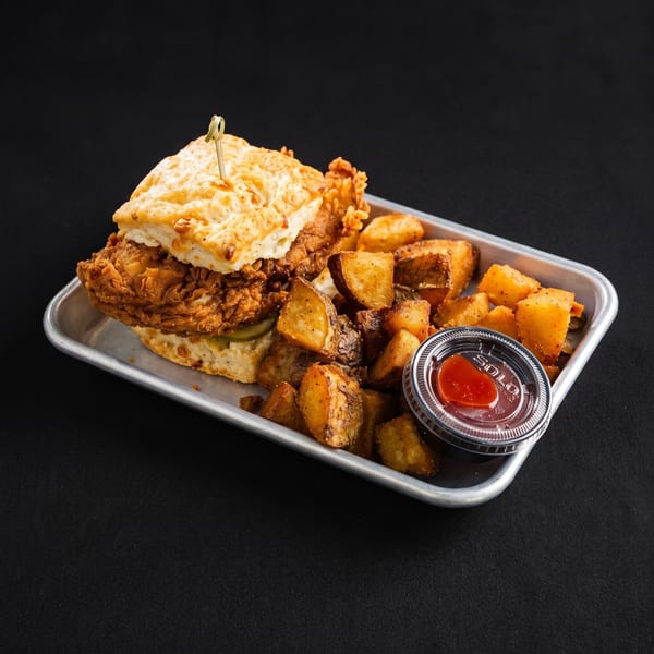 Crispy fried chicken biscuit sandwich with pickles, served with seasoned potato wedges and a side of sauce.