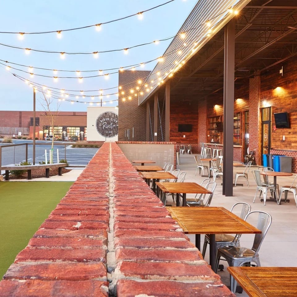 Outdoor patio of River City Roll with string lights, brick walls, wooden tables, and metal chairs.