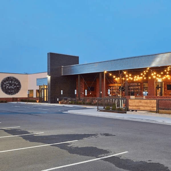 Exterior of River City Roll restaurant with a large parking lot and string lights on the patio.