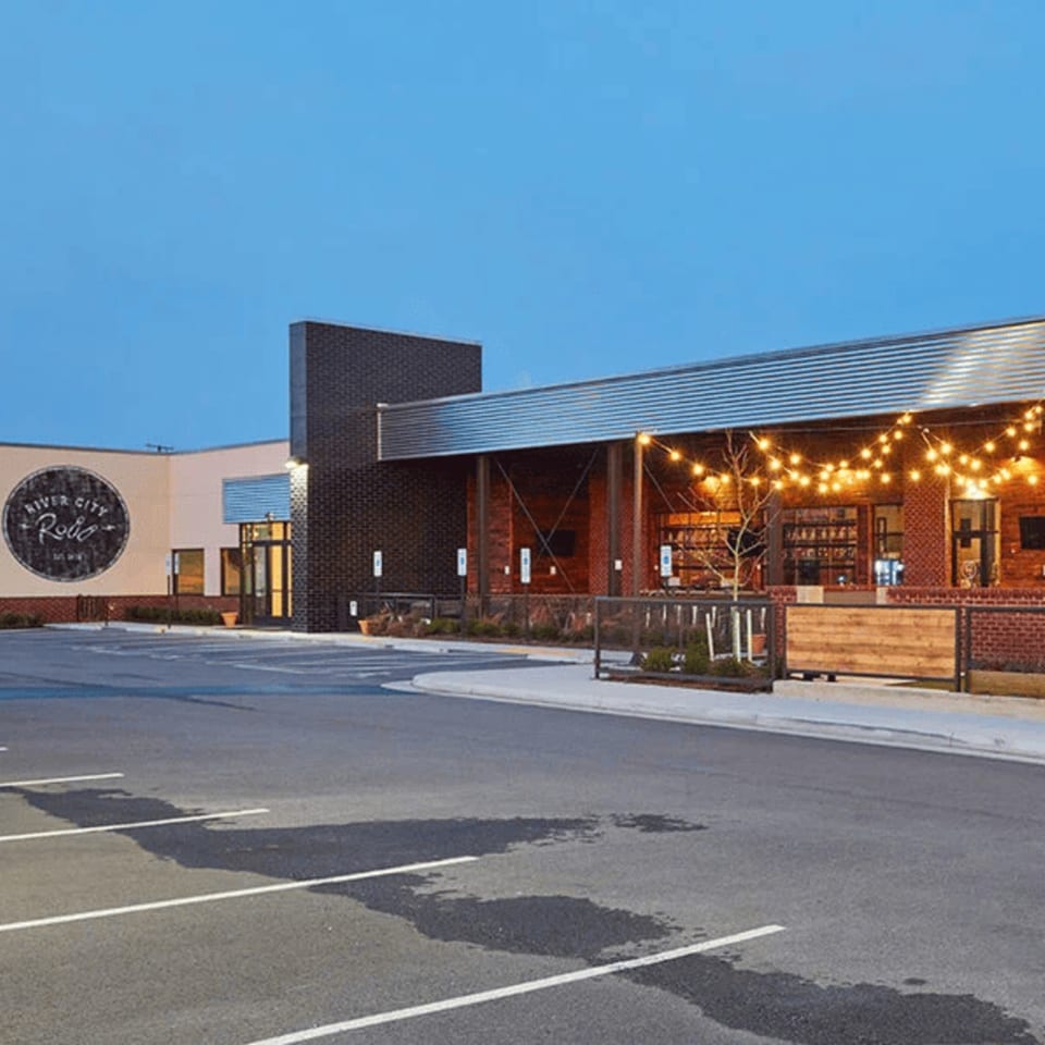 Exterior of River City Roll restaurant with a large parking lot and string lights on the patio.