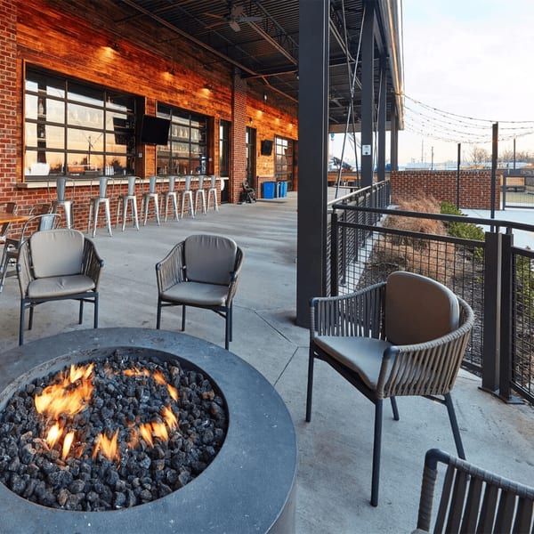 Outdoor patio with a lit fire pit surrounded by chairs, facing a brick building with a bar.