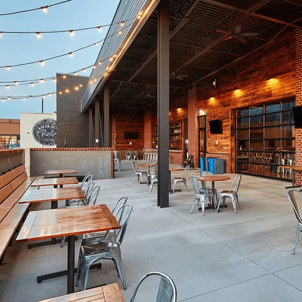 An outdoor patio with wooden tables, metal chairs, string lights, and a brick wall.