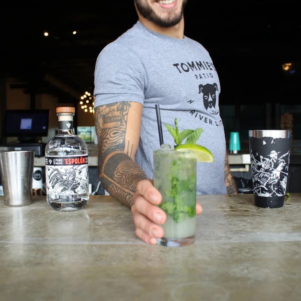 Bartender with tattooed arm serving a mojito cocktail, with tequila bottle and shaker on the bar.