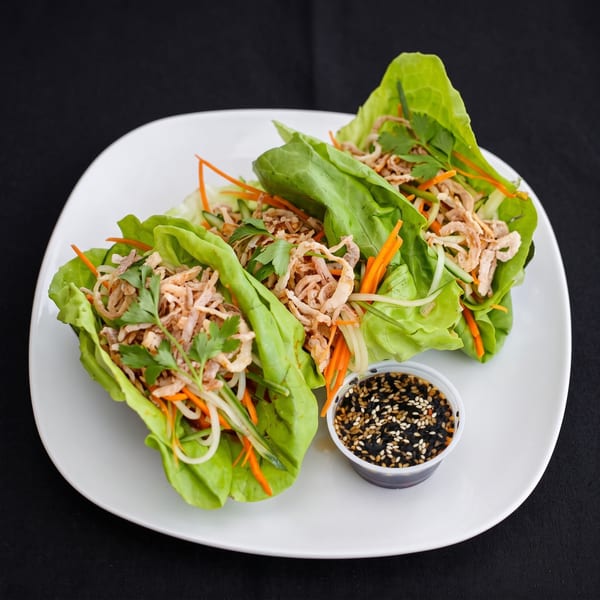 Three vibrant lettuce wraps filled with shredded chicken, carrots, and crispy fried onions, with a side of sesame sauce.