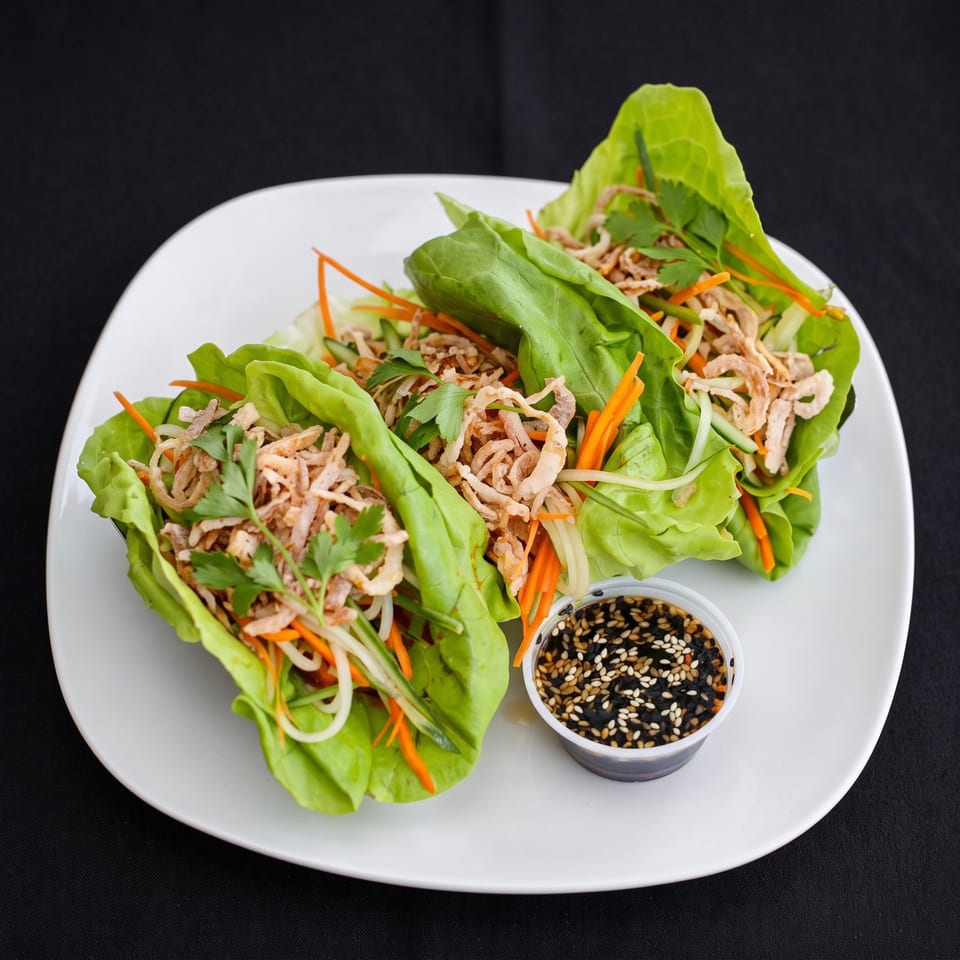Three vibrant lettuce wraps filled with shredded chicken, carrots, and crispy fried onions, with a side of sesame sauce.