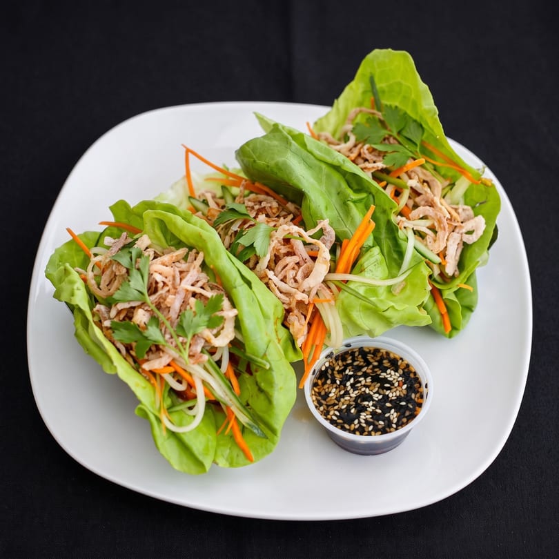 Three vibrant lettuce wraps filled with shredded chicken, carrots, and crispy fried onions, with a side of sesame sauce.