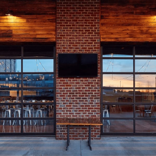 Exterior view of a restaurant with large windows, brick, and wood paneling, reflecting string lights and sunset.